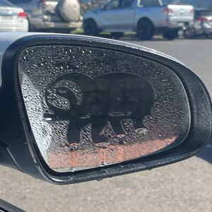 Könnte beinhalten: Autospiegel mit einem schwarzen Widder-Aufkleber. Der Spiegel ist mit Kondenswasser bedeckt. Der Hintergrund zeigt einen Parkplatz mit Autos und einen klaren blauen Himmel.