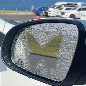 May include: A white car's rearview mirror with a clear butterfly sticker on it. The mirror is reflecting a blue sky and ocean with a few cars parked in the distance.