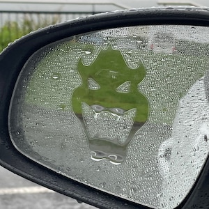 Puede incluir: Una pegatina de dragón verde en el espejo retrovisor de un coche. El espejo está cubierto de gotas de lluvia. La pegatina es de Cheeky Stickees, una pequeña empresa australiana orgullosa.