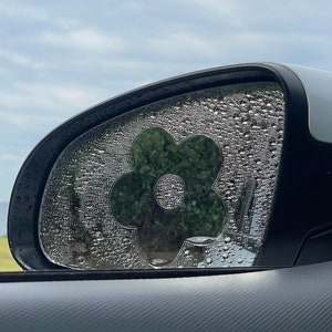 以下が含まれることがあります： 車のリアビューミラーに花柄のステッカーが貼られています。ミラーは水滴で覆われています。