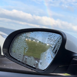 以下が含まれることがあります： 車のリアビューミラーに雨滴が落ちています。ミラーは、遠くの虹のある曇り空を映し出しています。ミラーには星型のステッカーが貼られています。ステッカーは透明で、白い輪郭があります。画像の右下には「CHEEKY STICKEES」というテキストが表示されています。