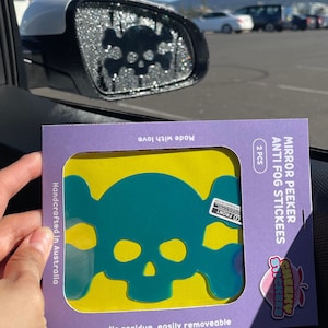 May include: Two teal skull and crossbones anti-fog stickers for car mirrors. The stickers are on a purple and yellow package with the text "Mirror Peeker Anti-Fog Stickees" and "Cheeky Stickees".