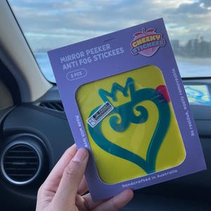 Könnte beinhalten: Eine Packung mit zwei gelben und grünen Anti-Beschlag-Aufklebern für Autospiegel. Die Aufkleber sind herzförmig mit einer Krone oben drauf. Die Verpackung trägt die Aufschrift "Mirror Peeker Anti Fog Stickees" und "Cheeky Stickees".
