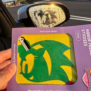 May include: A package of mirror peekers stickers featuring a green and yellow cartoon character. The package is purple with the words "MIRROR PEER STICKEES" and "2 PCS". The stickers are "No residue, easily removeable".