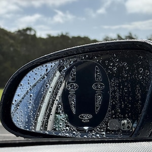 Könnte beinhalten: Ein Seitenspiegel eines Autos mit Regentropfen auf der Oberfläche. Ein schwarz-weißer Aufkleber mit einem Comic-Gesicht befindet sich auf dem Spiegel.