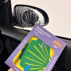 May include: A purple cardboard packaging containing two green and yellow shell-shaped stickers. The packaging features the text MIRROR PEEKER STICKEES, 2 PCS, CHEEKY STICKERS, Made with love, and Handcrafted, easily removeable.