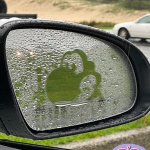 Könnte beinhalten: Ein klarer Autospiegel mit einem grün-weißen Aufkleber eines Cartoon-Geistes mit einem breiten Lächeln. Der Aufkleber befindet sich am Beifahrer-Spiegel eines Autos. Der Spiegel ist mit Regentropfen bedeckt.
