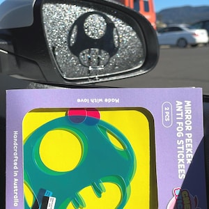 Op de afbeelding: Twee groene en blauwe anti-condens stickers in de vorm van een paddenstoel voor autoruiten. De stickers zijn verpakt op een paarse en gele kaart met de tekst "Mirror Peeker Anti Fog Stickees" en "Cheeky Stickees".