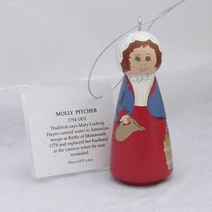 May include: Wooden doll ornament depicting Molly Pitcher, a historical figure who carried water to American troops during the Battle of Monmouth in 1778. The doll is painted in red, blue, and white, and holds a brown pitcher in her right hand. The doll is wearing a white bonnet, a blue jacket, and a red skirt. The ornament is labeled "Molly Pitcher 1754-1832".