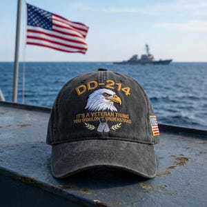 May include: A dark gray baseball cap with the text "DD-214" and an eagle graphic. The cap also features the phrase "It's a Veteran Thing You Wouldn't Understand" and a small American flag patch. The background includes the American flag, the ocean, and a ship.