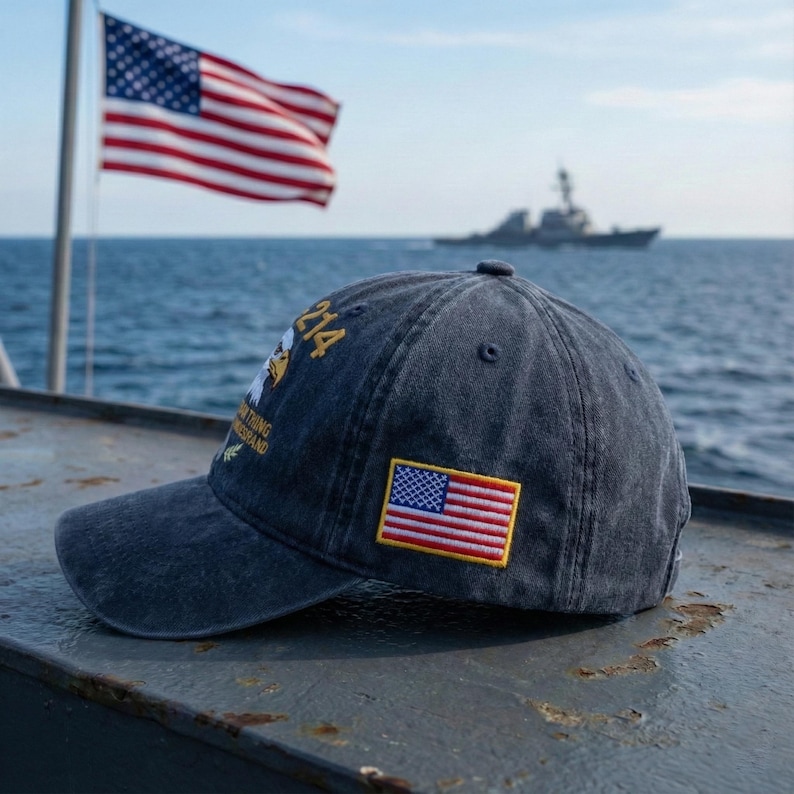 May include: A navy blue baseball cap with a small American flag patch and text. The cap has a curved brim and is set against a backdrop of the ocean, an American flag, and a ship.