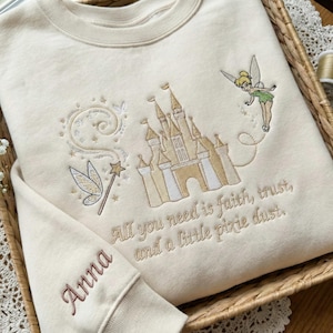 May include: Cream-coloured sweatshirt with embroidered design featuring a castle, fairy, and text "All you need is faith, trust, and a little pixie dust." The sleeve has the name "Anna" embroidered in a matching colour.