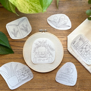Puede incluir: Una colección de pegatinas de arte lineal y un bastidor de bordado con diseños botánicos y temáticos de libros. Las pegatinas incluyen imágenes de un libro abierto, setas, una pila de libros con una planta, una cómoda, una taza y una planta en maceta.