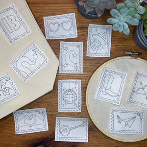 May include: Embroidered patches in postage stamp designs, featuring a variety of images such as a snake, a heart-shaped hand gesture, a disco ball, and a cowboy boot. Some patches are attached to a canvas bag and an embroidery hoop.