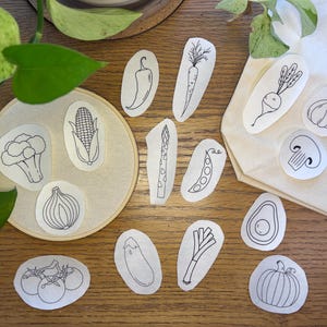 Puede incluir: Un conjunto de 13 círculos blancos con dibujos de línea negra de varias verduras y frutas, incluyendo un aguacate, una calabaza, una mazorca de maíz, un hongo, una cebolla, una vaina de guisantes, una berenjena, un puerro, un brócoli, un chile, espárragos, una zanahoria y un nabo.