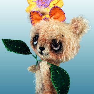 May include: A small, furry, brown teddy bear wearing a yellow, orange and purple flower on its head. The bear is holding a green leaf in its paws.