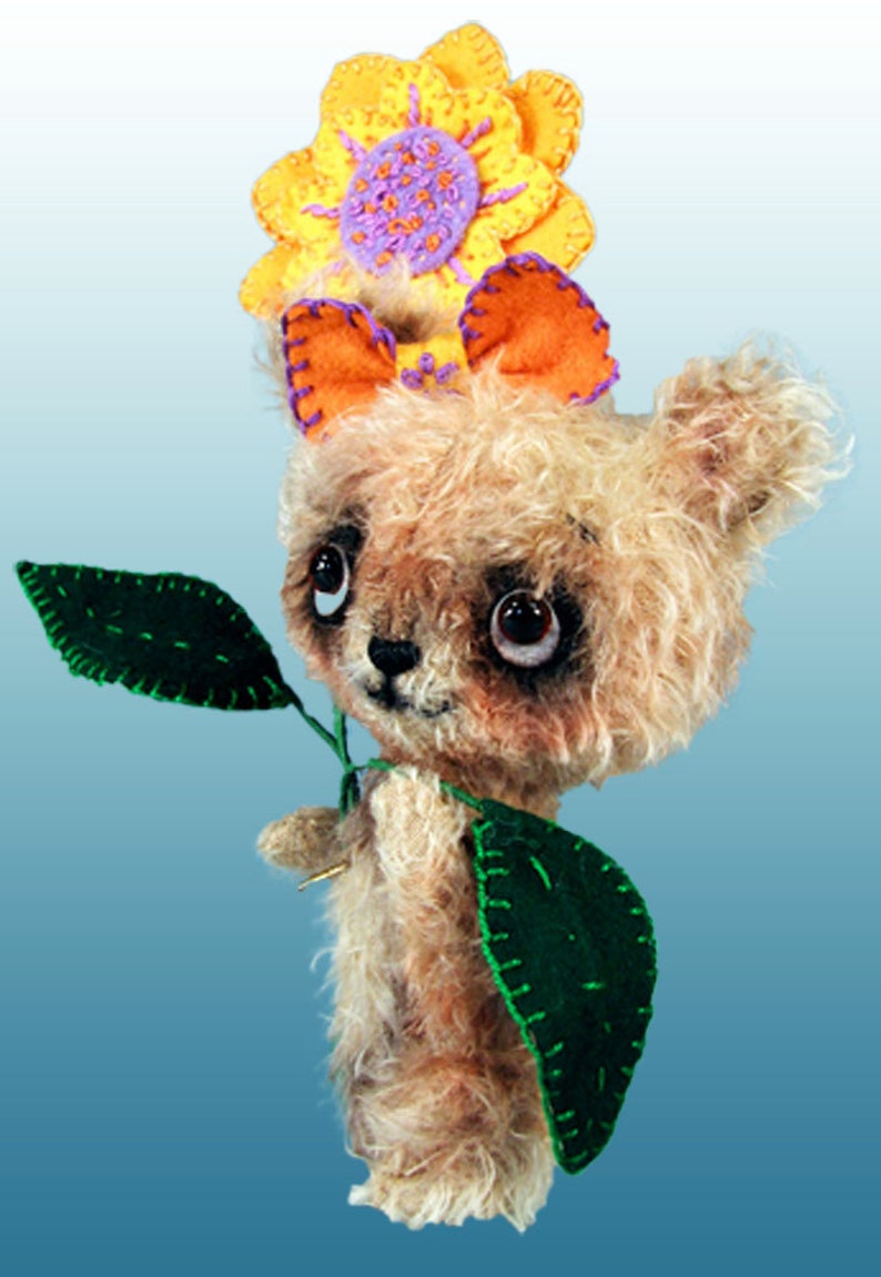 May include: A small, furry, brown teddy bear wearing a yellow, orange and purple flower on its head. The bear is holding a green leaf in its paws.