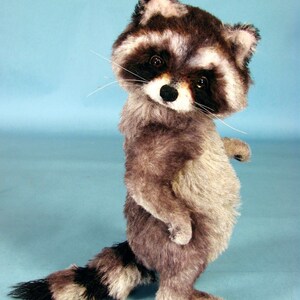 May include: A plush raccoon toy standing on a light blue background. The raccoon has a black and white face with a black nose and black eyes. The raccoon is wearing a gray and white fur coat.