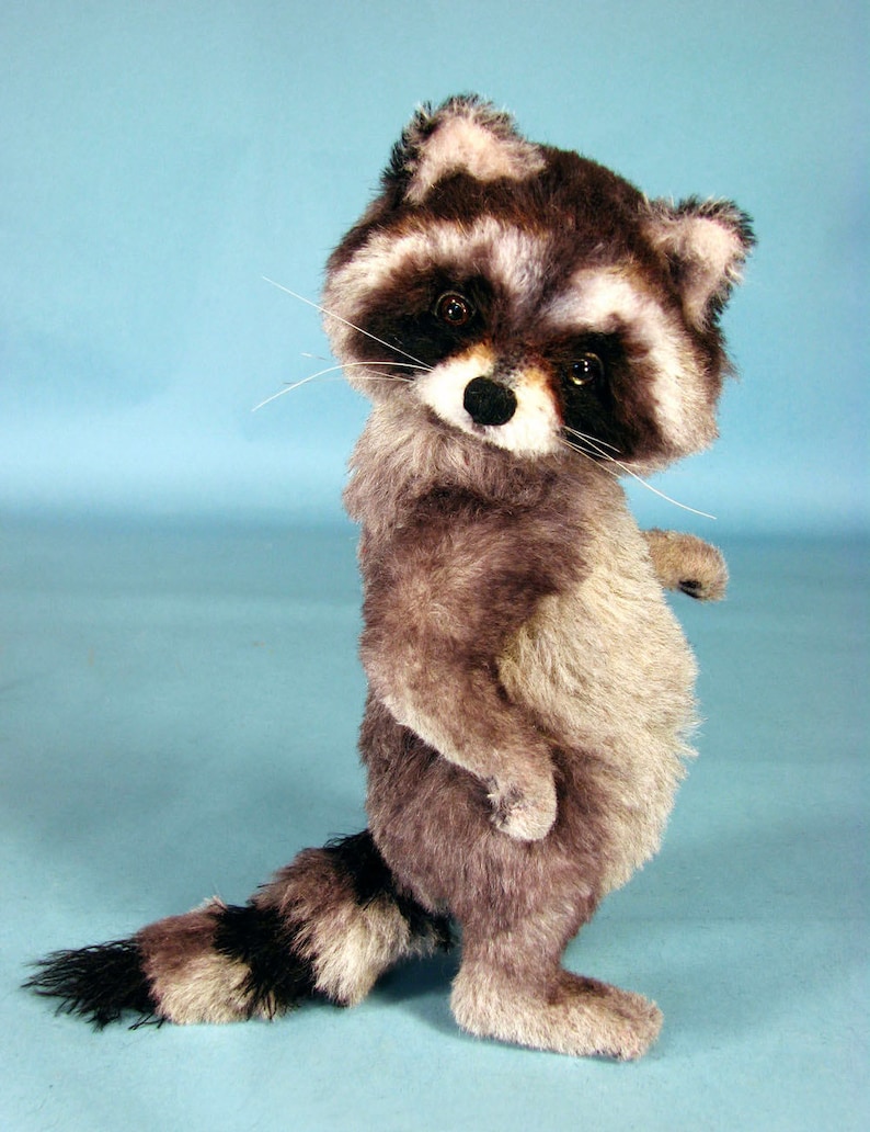 May include: A plush raccoon toy standing on a light blue background. The raccoon has a black and white face with a black nose and black eyes. The raccoon is wearing a gray and white fur coat.