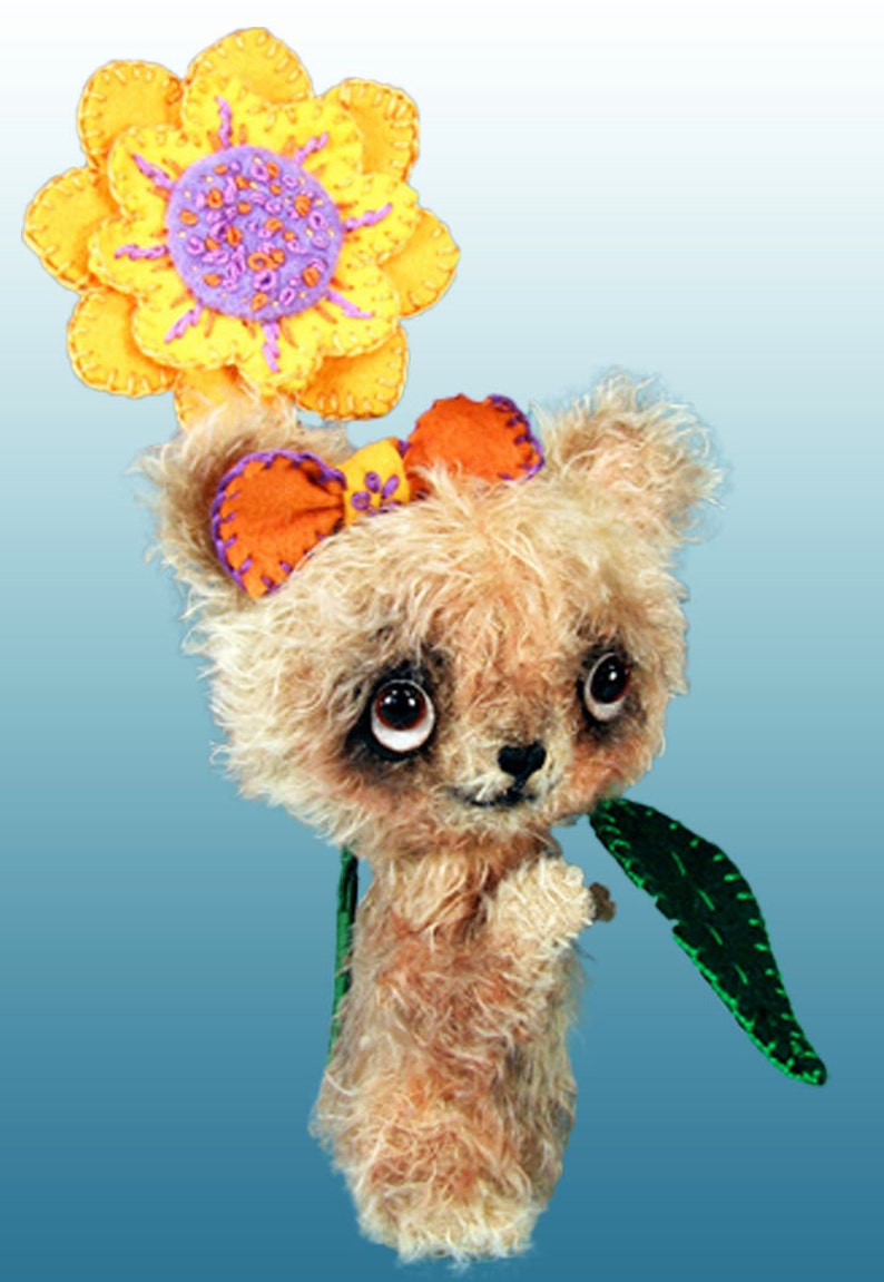 May include: A small, furry, brown teddy bear wearing a yellow flower on its head and an orange bow. The bear is holding a green leaf in its paw.