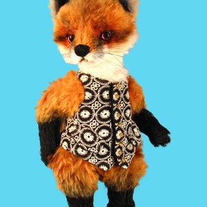 May include: A plush fox toy wearing a black and white patterned vest with gold buttons. The fox has orange fur, black eyes, and a black nose.
