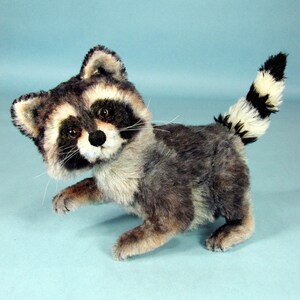 May include: A plush raccoon toy with a black and white striped tail, gray and black fur, and black eyes. The toy is standing on a light blue background.