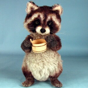 May include: A plush raccoon toy holding a small wooden bucket. The raccoon has a gray and brown fur with black eyes and a black nose. The toy is standing on a light blue background.