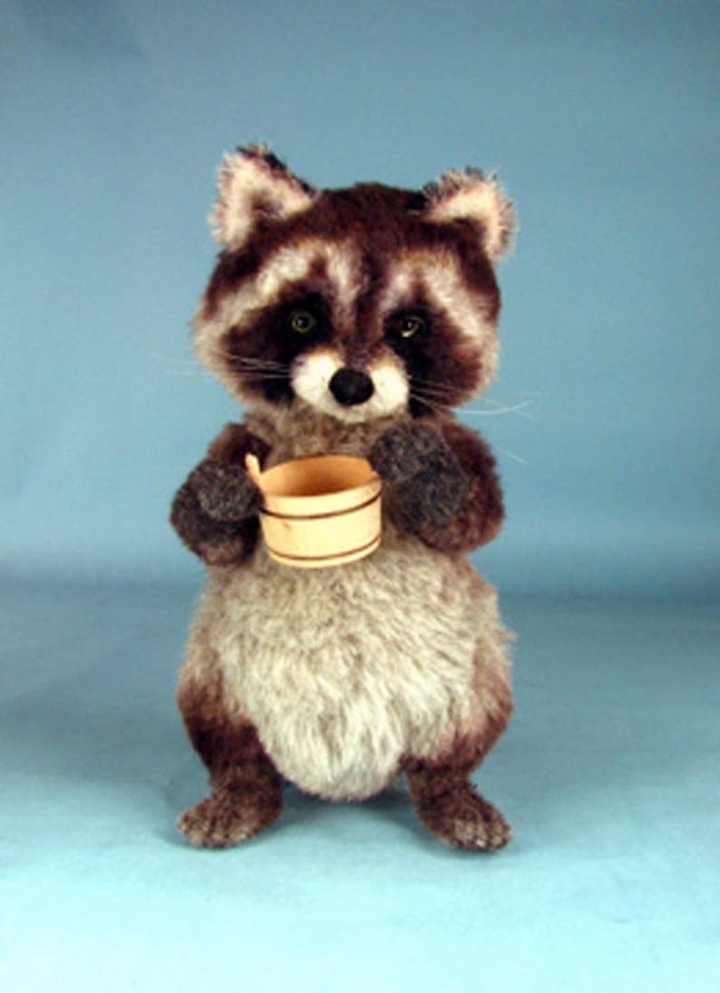 May include: A plush raccoon toy holding a small wooden bucket. The raccoon has a gray and brown fur with black eyes and a black nose. The toy is standing on a light blue background.