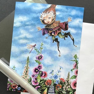 May include: A whimsical art print featuring a fairy-like figure with a pointed hat, floating in a blue sky with clouds. The illustration includes a vibrant flower garden with pink, red, and blue blooms, and a dragonfly. A silver pen is in the foreground.