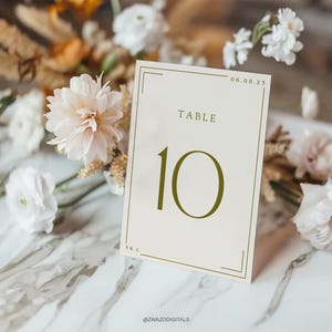 May include: A white table number card with a gold border and the number 10 in a gold font. The text "Table" is above the number and the date "06.08.25" is in the top right corner. The card is sitting on a marble surface with flowers.