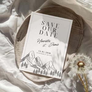 Op de afbeelding: Save the date-kaart met een illustratie van een bergketen en een bos. De kaart zegt "Save Our Date" met de namen "Henrietta & Jamie" en de datum "14.05.2030". Het adres "123 Anywhere St., Any City" is ook inbegrepen. De kaart ligt op een lichtbruin oppervlak.