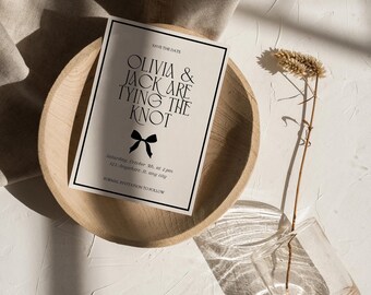 May include: A wedding invitation with the text "Olivia & Jack Are Tying The Knot" in a black font, featuring a bow graphic. The invitation is set in a wooden bowl with a dried plant in a glass vase. The text "Save The Date" is at the top.