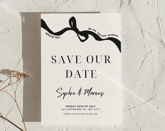 May include: A white save the date card with a black ribbon bow and the text "WE ARE GETTING MARRIED."  The text "25TH OF JULY" is on the ribbon. The text "SAVE OUR DATE" is in large, bold letters. The names "Sophie & Marcus" are written in cursive. The date "FRIDAY 25TH OF JULY" and the address "123 ANYWHERE ST. ANY CITY." are printed below the names. The text "FORMAL INVITATION TO FOLLOW" is printed below the address. The text "@ZWAZODIGITALS" is printed at the bottom of the card.
