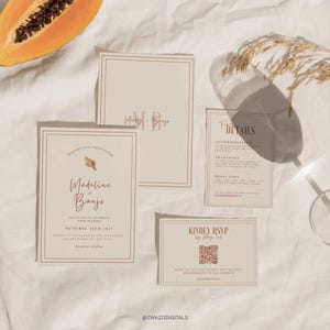 May include: A collection of beige wedding invitation cards with a minimalist design. The cards feature brown script text, including names, dates, and RSVP details. The cards are arranged on a white surface with a papaya half and dried grass.