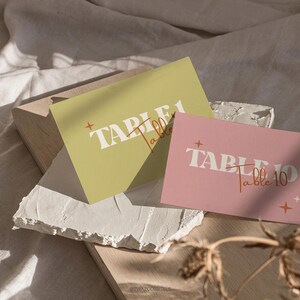 Puede incluir: Dos tarjetas de número de mesa, una amarilla y otra rosa, con el texto "TABLE 1" y "TABLE 10" en blanco. El texto "Table 1" está escrito en cursiva naranja. El texto "Table 10" está escrito en cursiva naranja. Ambas tarjetas tienen un pequeño diseño de estrella en la esquina superior derecha.