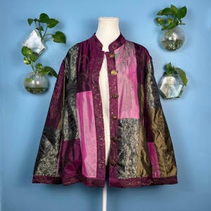 May include: A patchwork jacket in shades of purple, pink, and olive green. The jacket features a mandarin collar, gold-colored buttons, and ornate embroidery. The jacket is displayed on a mannequin.