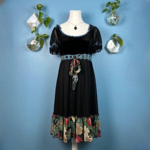 Peut inclure: Une robe en velours noir avec une bordure en crochet bleu et une jupe à imprimé floral. La robe a un col rond et des manches courtes avec des pompons.