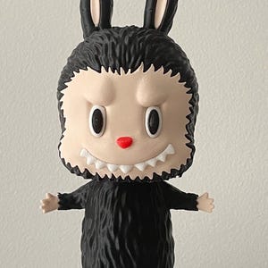 May include: A black and beige figurine of a creature with rabbit ears, a furry body, and a menacing expression. The figurine has a red nose, white teeth, and outstretched arms. The toy is standing on a white surface.