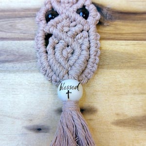 Op de afbeelding: Een handgemaakte macramé uil sleutelhanger in een lichtbruine kleur. De uil heeft zwarte kraalogen en een hartvormig ontwerp in het midden. Een houten kraal met het woord "blessed" en een kruis is bevestigd aan een kwast.
