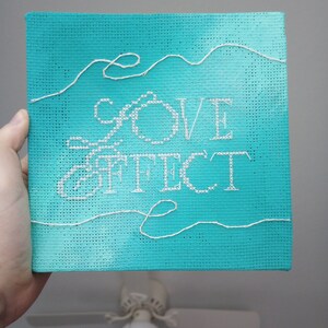 May include: A square, turquoise canvas with the words "LOVE EFFECT" stitched in white thread. White thread accents the top edge. The canvas is held by a hand.