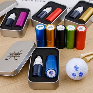 May include: A collection of golf-themed items, including ink bottles, cylindrical stamps, and a golf ball. The stamps come in various colors, and the golf ball features a blue and white design. The items are presented in small metal tins.