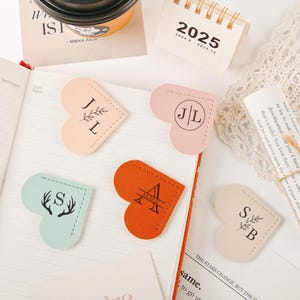 Puede incluir: Cuatro marcapáginas de cuero en forma de corazón con diferentes colores e iniciales personalizadas. Los marcapáginas son de color rosa claro, azul claro, naranja y beige. Las iniciales son "JL", "S", "ALICE" y "SB".