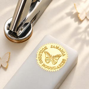 May include: A silver metal embosser tool positioned next to a white rectangular block featuring a gold circular seal. The seal has a serrated edge and displays a butterfly graphic with the text JAMAICA BUILDING BOUNDARIES embossed on it.