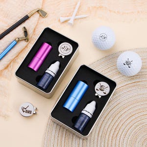 May include: Golf gift set with two metal boxes, each containing a cylindrical item, a small bottle, and a ball marker. Two golf balls and white tees are also present. The boxes have black foam inserts. The golf clubs are gold and black.
