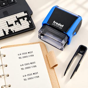 May include: A blue and black Trodat Printy 4911 typo self-inking stamp, a black character set tray, and black plastic tweezers. A tan notebook displays the stamped text A-2533 WEST and TEL: 2065/1709 repeated three times on white paper.