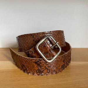 Op de afbeelding: Een bruine slangenleren riem met een zilverkleurige vierkante gesp. De riem is opgerold op een houten oppervlak, waarbij het textuurpatroon en de kleurvariaties te zien zijn. De gesp is gecentreerd.