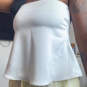 May include: A white strapless top with a peplum hem. The top is made of a smooth, silky fabric. The model is wearing yellow and white striped trousers. A gold necklace is visible.