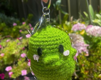 May include: Green crocheted ghost keychain with a cute face. The ghost has black eyes and pink cheeks. The keychain is attached to a silver key ring.