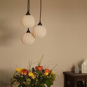 Pu&ograve; includere: Tre lampade a sospensione con paralumi in vetro bianco a spirale sono appese a un apparecchio a soffitto bianco. Sotto, un vaso bianco contiene una colorata composizione floreale di fiori gialli, arancioni e rossi.