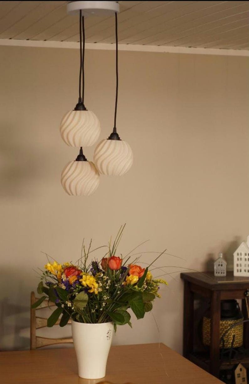 Pu&ograve; includere: Tre lampade a sospensione con paralumi in vetro bianco a spirale sono appese a un apparecchio a soffitto bianco. Sotto, un vaso bianco contiene una colorata composizione floreale di fiori gialli, arancioni e rossi.